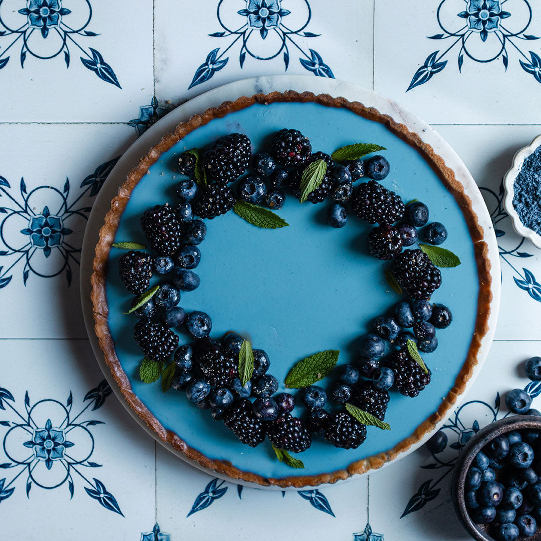 A tart with a smooth blue filling is decorated with blackberries, blueberries, and mint leaves, arranged in a wreath on top. The tart sits on a white and blue patterned tile surface.